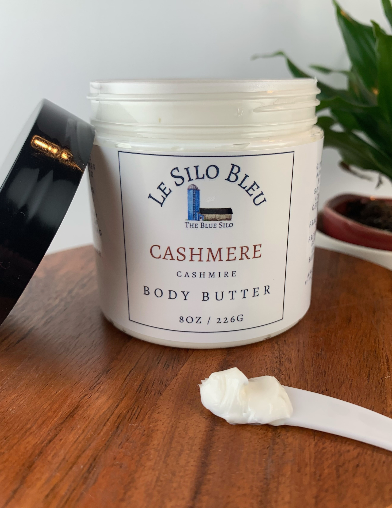 An open jar of Body Butter on a wooded table with it's black lid leaning to the left of the jar and with a small white plastic spoon scooped with the lotion on it. There is a green plant in a brown pot in the background. 