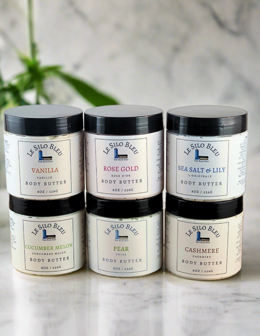 six jars of body butter in various scents sitting on top of a marble table with a white background.  The scents shown are in various colors according to their scent.