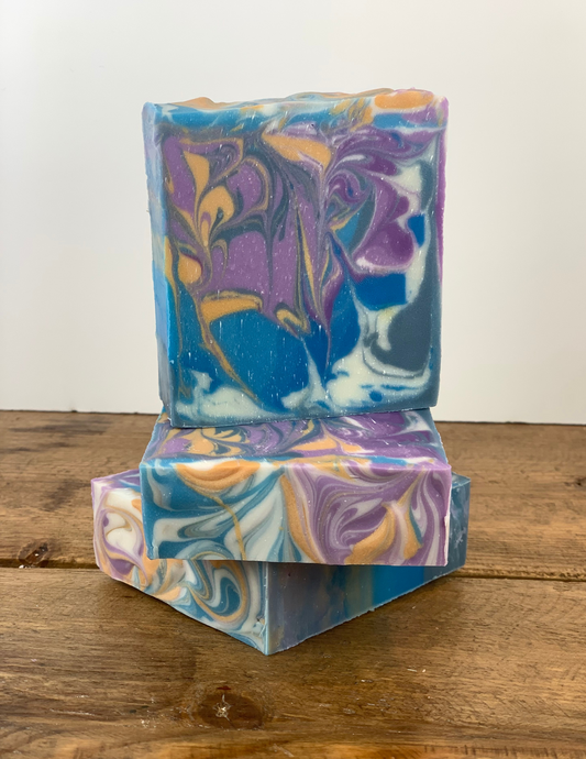 Blue, purple, gold and white swirled soap stacked  on top of a wooden table with a white background. 