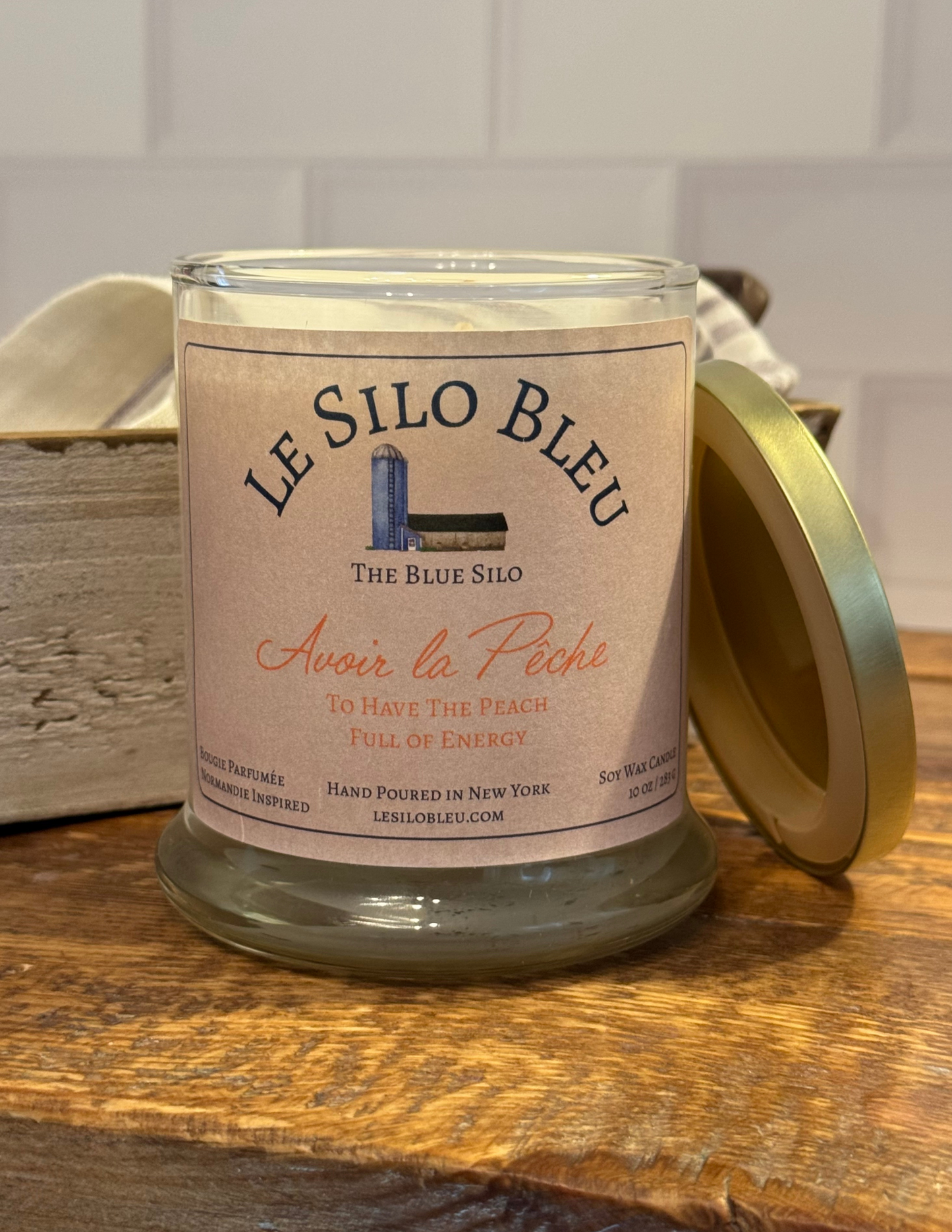 Avoir la Peche, a peach scented candle with gold lid to the side. Candle is sitting on top of a wood table with an antique box and linen cloth in the background.