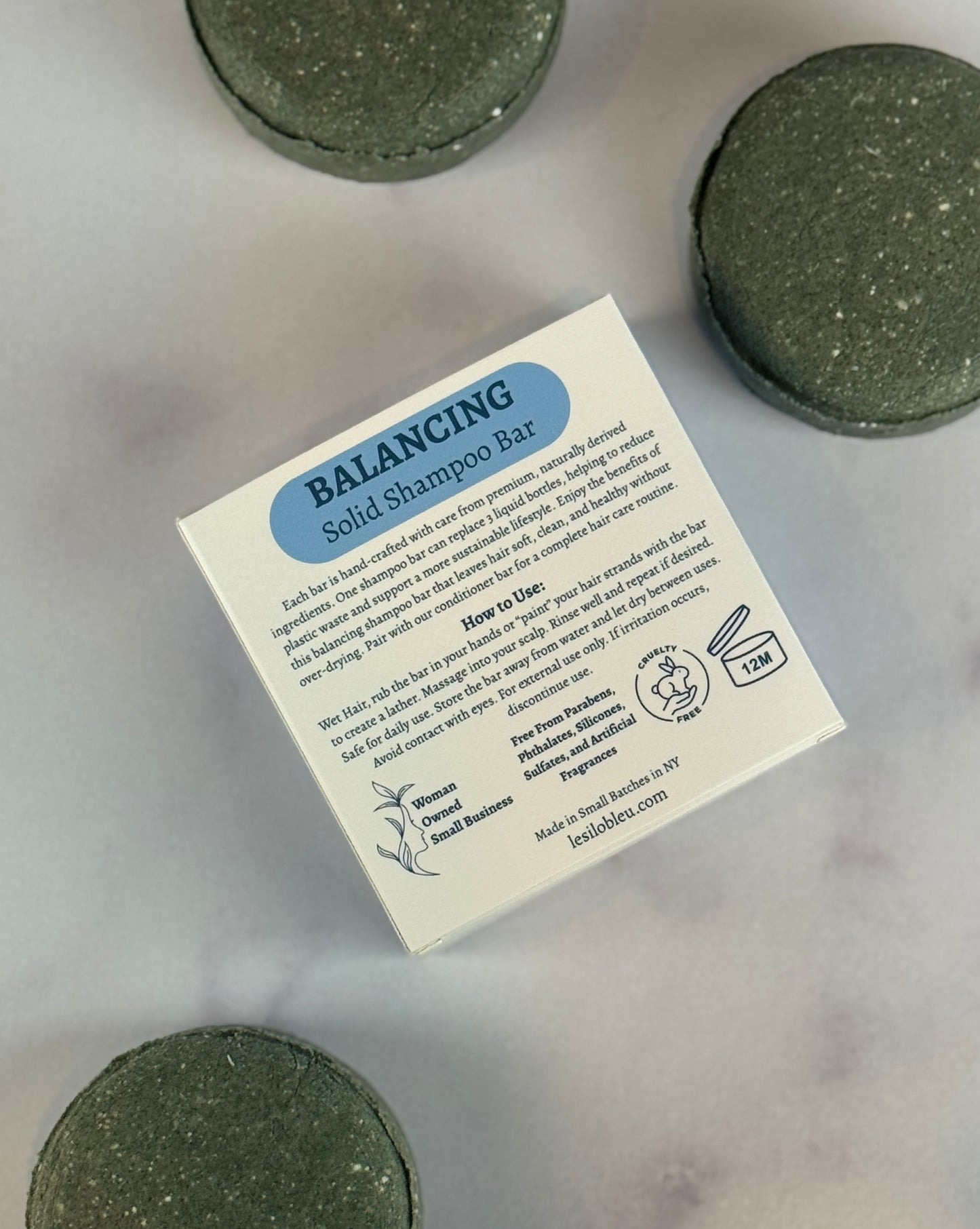 Three dark green scattered Balancing Solid shampoo bars with a back of the box label on a marble surface