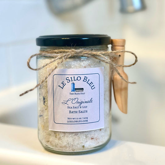 A jar of Sea Salt and Lily Bath salts sits on the edge of the bathtub. Details shows the jar tied with twine and a small wooden scoop is attached. Tub Faucet is blurred in the backgound.