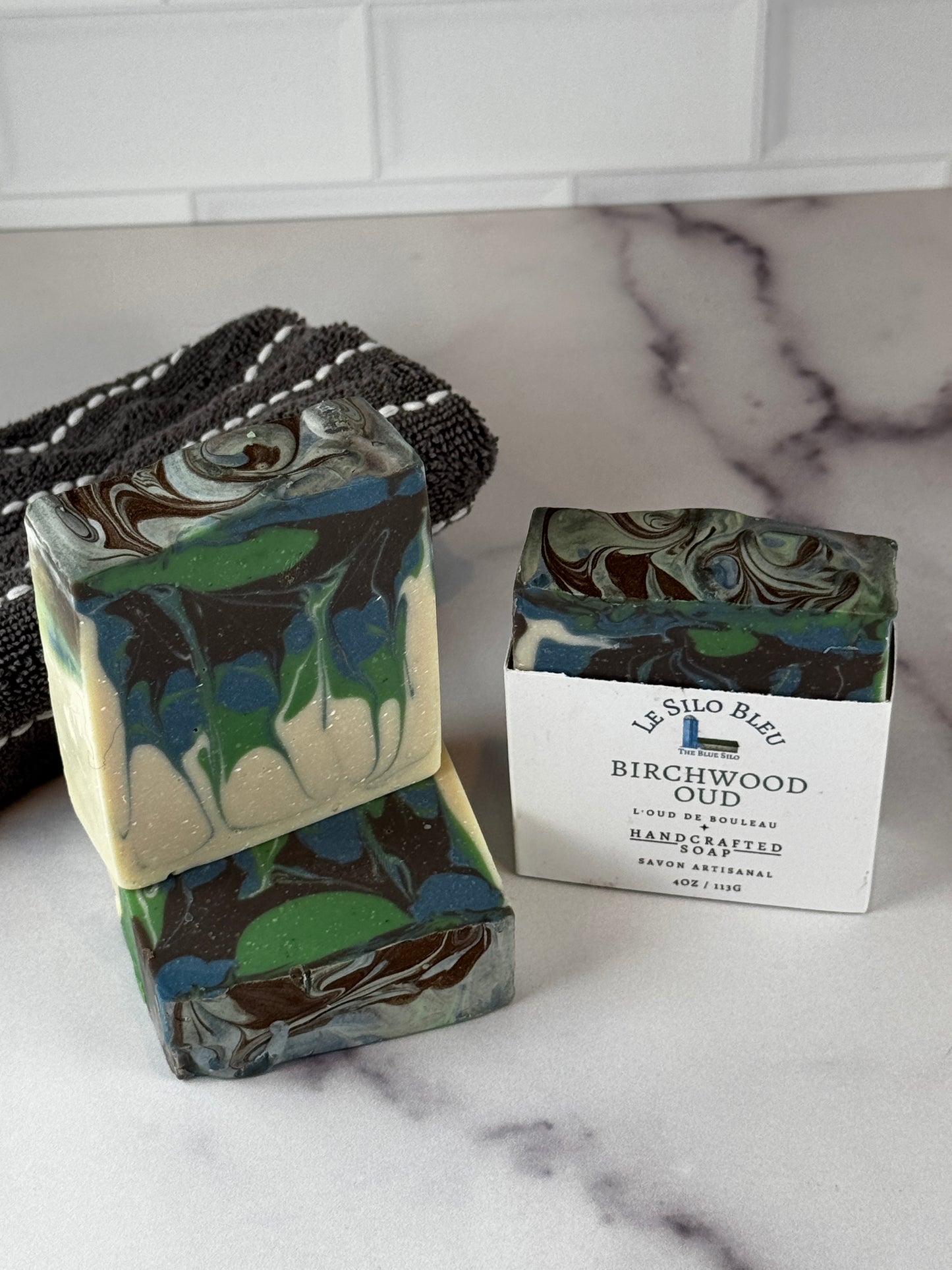 Two handmade soap bars with a marbled swirl pattern in green, blue, brown, and cream, displayed on a white marble surface next to a black-and-white striped cloth. One bar is standing upright with a white paper label that reads 'Silo Blue Birchwood'.