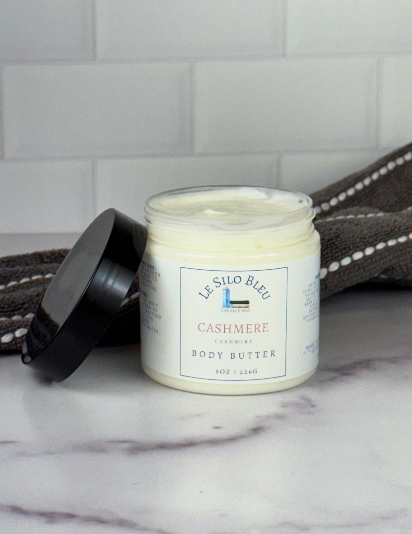 An open jar of Cashmere scented Body Butter with a black lid leaning against the jar sitting on top of a white marble table with a grey and white stripped towel in the background. The label shows the logo and the name of the scent.