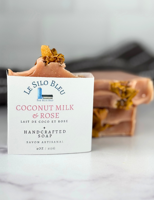 One labeled bar and two naked bars of pink soap with rose petals on top sitting on a white marble table with a grey and white stripped towel in the background.