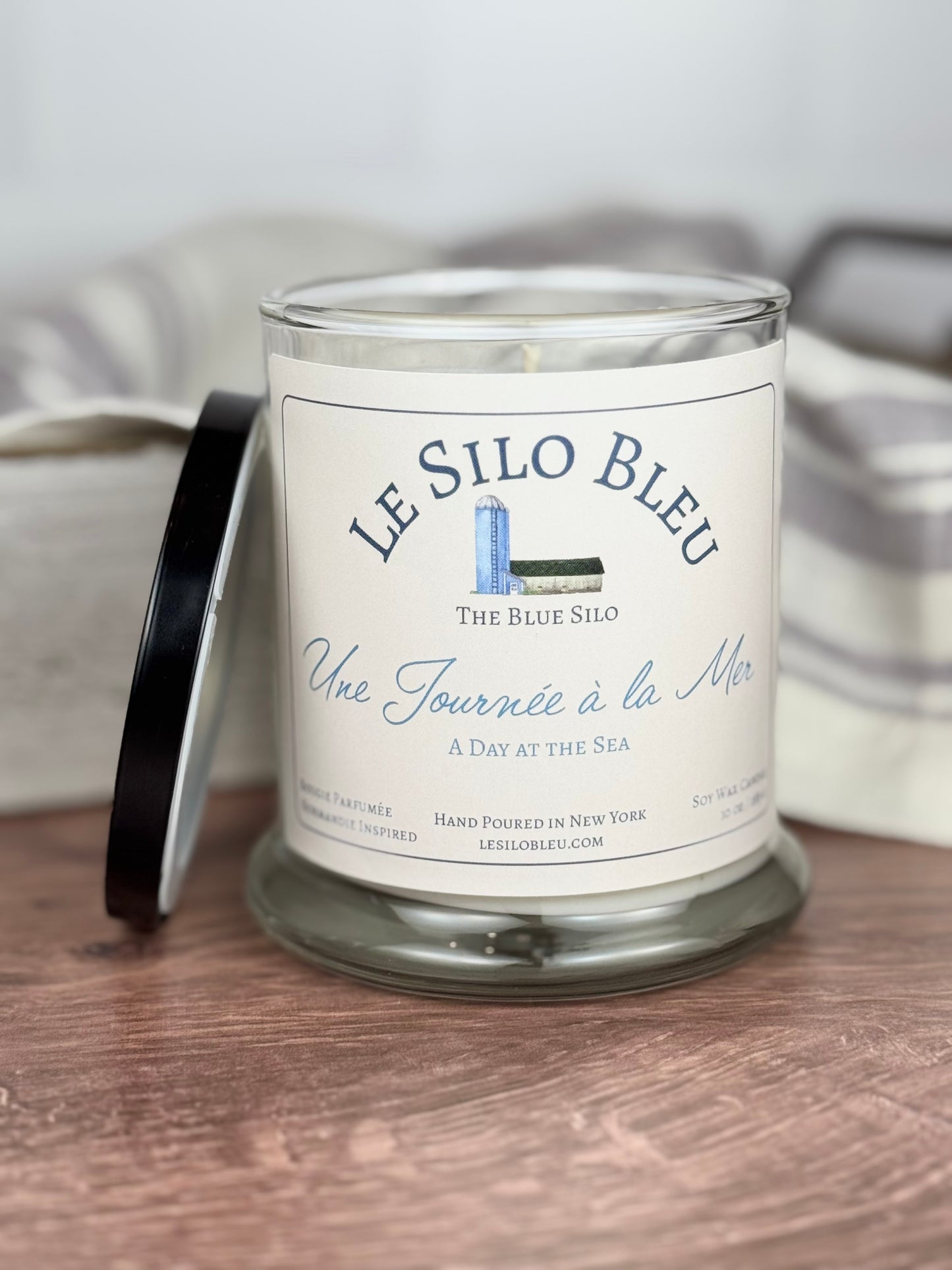 A large handpoured unlit soy wax candle in a glass jar labeled "Une Journee a la Mer- Day at the Sea" sitting on a wooden table top with a black lid next to the jar. In the background there is a white linen towel with lavender stripes in an antique box with a metal handle.