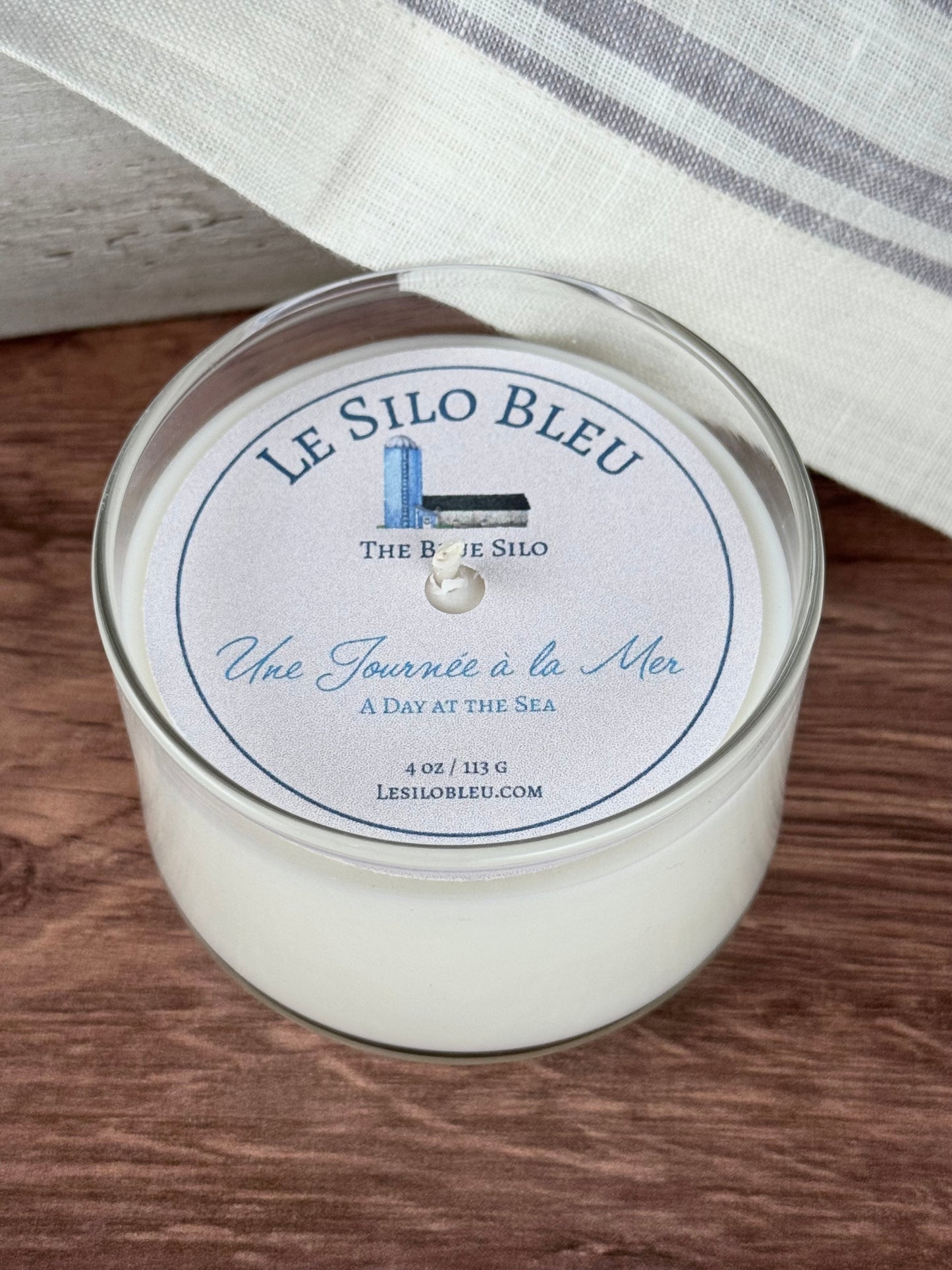 A small handpoured unlit soy wax candle in a glass jar labeled "Une Journee a la Mer- Day at the Sea" sitting on a wooden table top with a black lid on the jar. In the background there is a white linen towel with lavender stripes in an antique box.