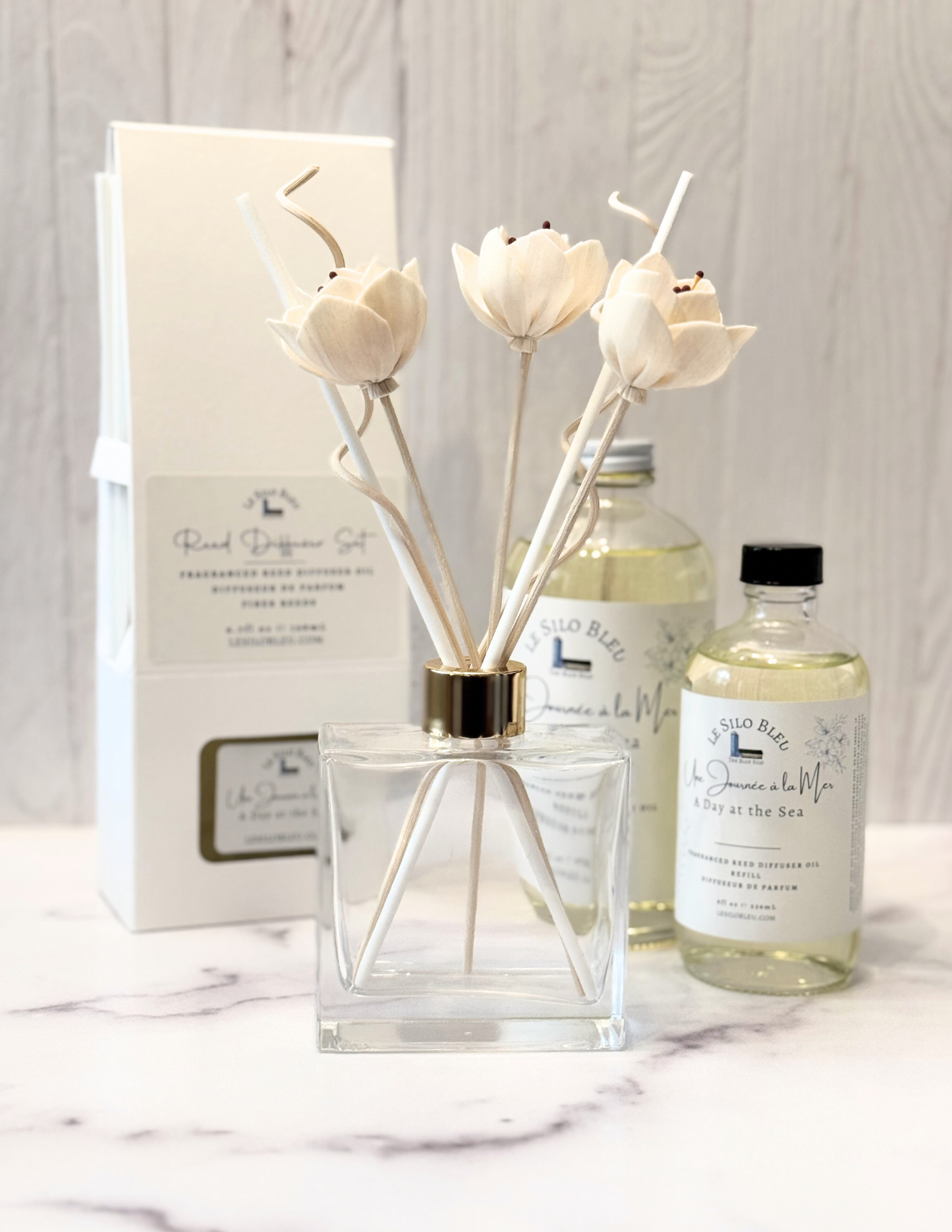Day at the Sea Scented Reed Diffuser set and collection showcasing the optional floral reeds along with the included white reeds and additional for purchase bottles of 8oz or 16oz fragrance refills. The whole scent collection is sitting on a white marble table with a white wood background.