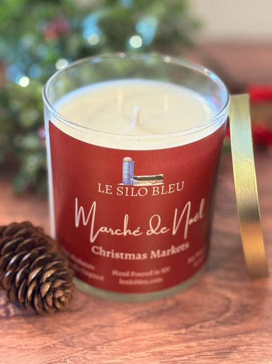Candle with a red label featuring 'Le Silo Bleu' on a wooden surface with a blurred Christmas tree background.