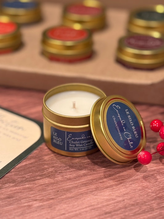 Candle in a gold tin with a blue label on a wooden surface, surrounded by other candles.