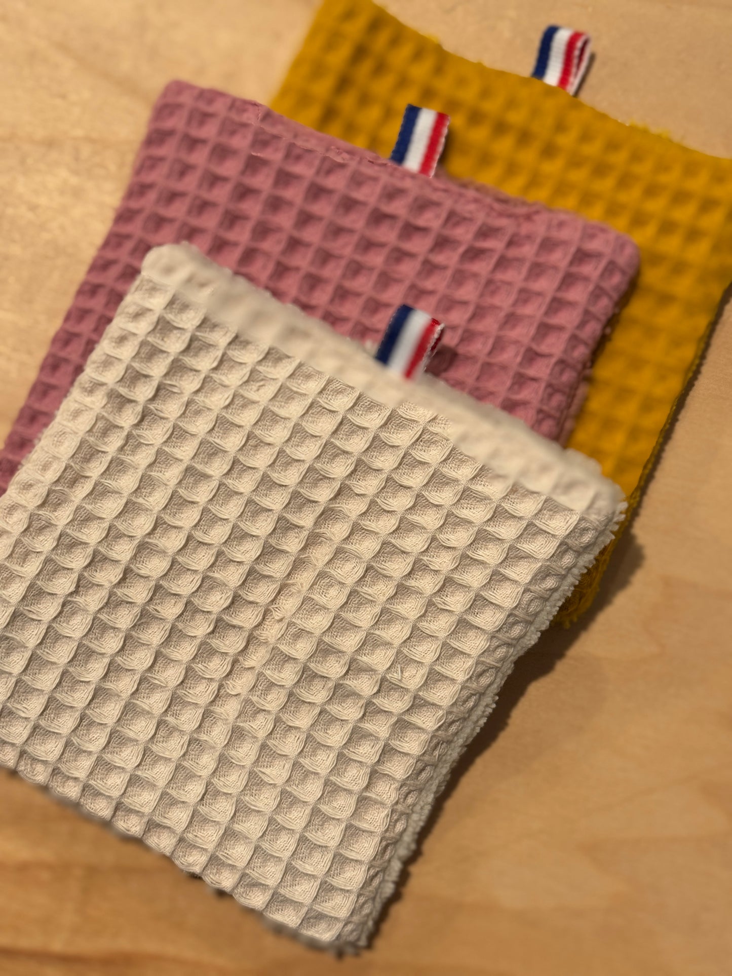 Three textured cotton cosmetic pads in pink, beige, and yellow on a wooden surface.