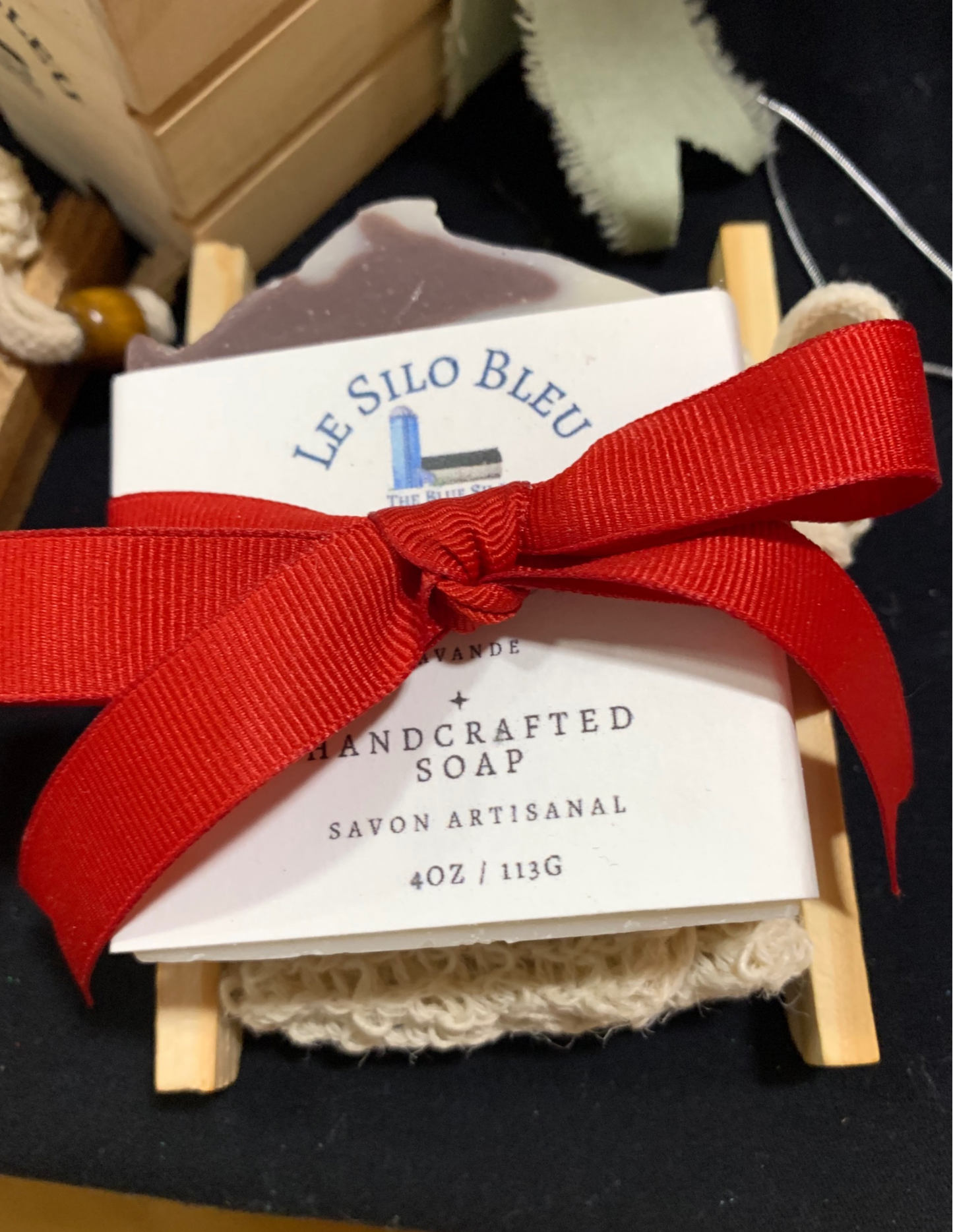 A bar of our handmade soap with a soap saver bag all tied together with a red ribbon on a wooden bamboo soap tray. This gift set is on top of a black tablecloth with a crate of soap and another gift bundle visible in the corners of this picture.