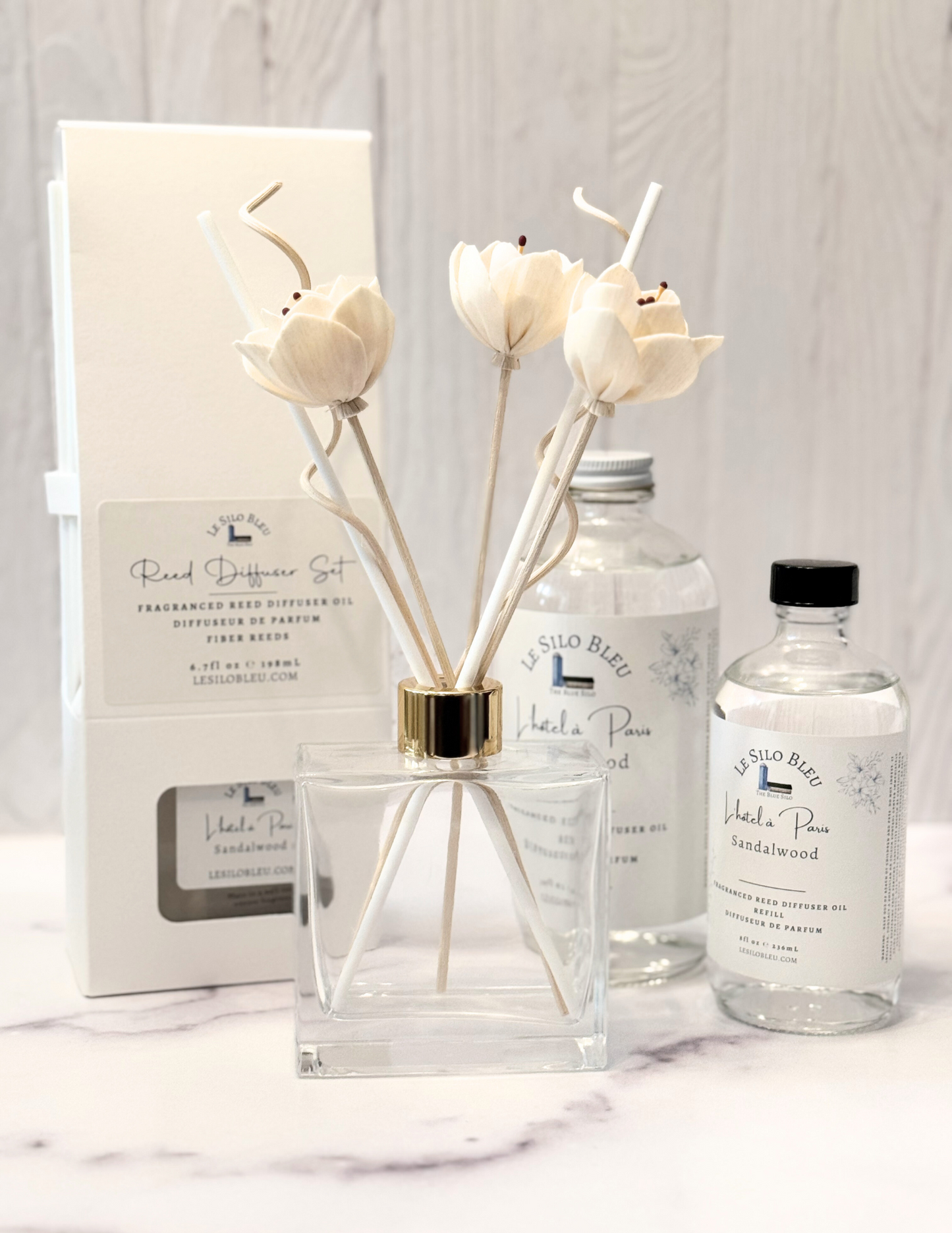 Hotel in Paris Reed Diffuser set and collection of for additional purchase refills and optional botanical floral reeds are all sitting on a white marble table with a white wood background.