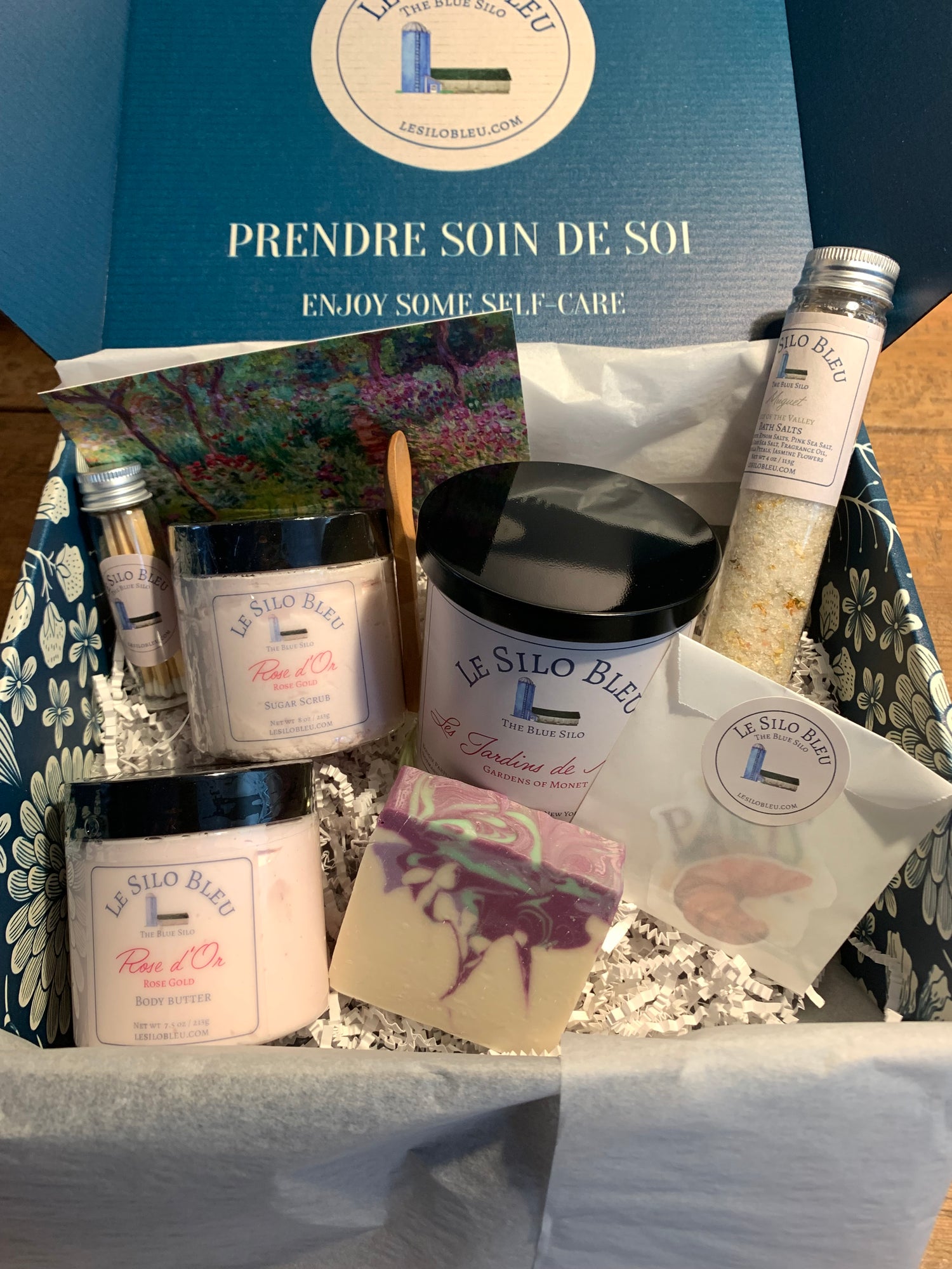 Le Silo Bleu Gift box with soap, body butter, sugar scrub, a large 10oz candle, matches in a tube and a tube of bath salts all packaged in a premium branded box with blue and white flowers printed on it. Also included is a small wooden scoop for your bath products, a postcard and some France inspired stickers.