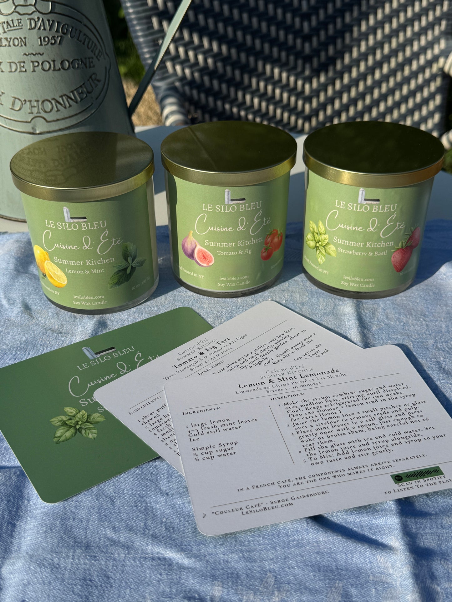 Three kitchen collection candles with gold lids on a blue surface, accompanied by recipe cards.