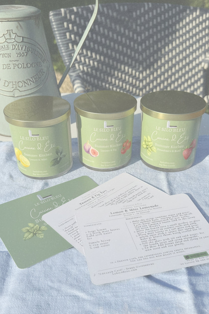 Three kitchen collection candles with gold lids on a blue surface, accompanied by recipe cards.