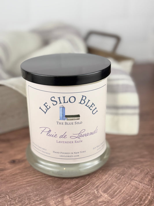 A handpoured unlit soy wax candle in a glass jar labeled "Pluie de Lavender - Lavender Rain" sitting on a wooden table top with a black lid on  the jar. In the background  there is a white linen towel with lavender stripes in an antique box with a metal handle.