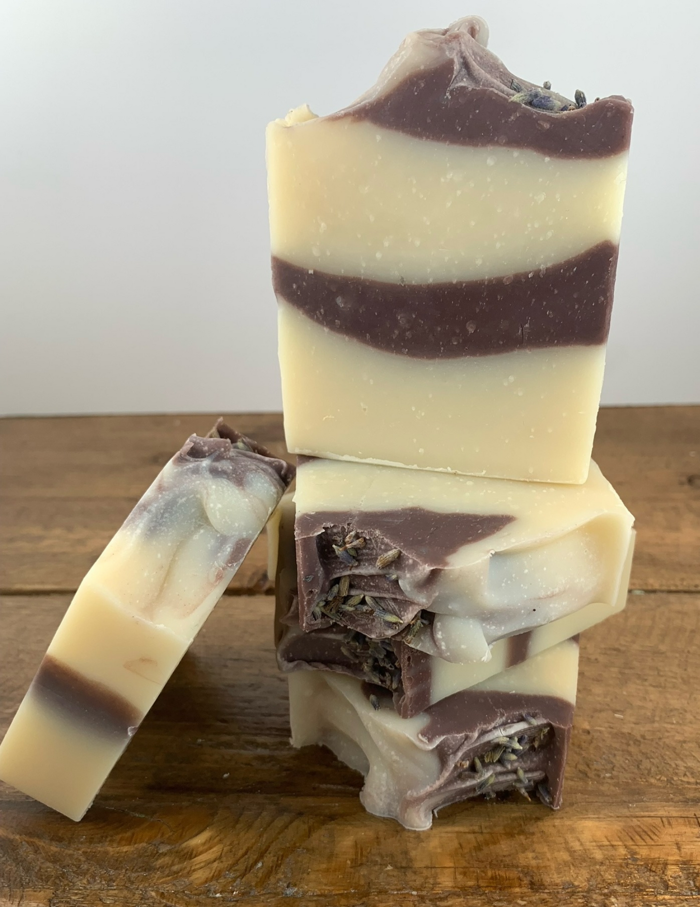 Stack of Lavender Soaps on a wooden table with a white background. Another bar of soap leans against the stack. The soap is a creamy color with a purple swirl from the Brazillian Purple Clay.