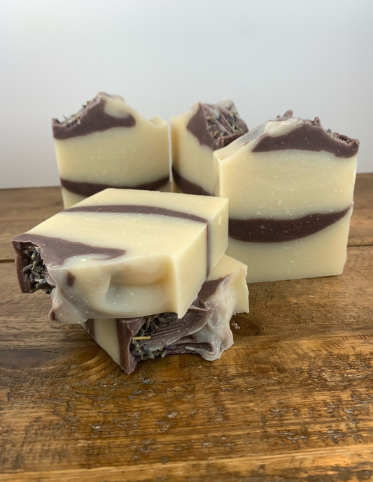 This handcrafted, palm free, cold-processed soap is made with Purple Brazilian Clay, and essential oil for an all natural bar. Bars of soap on a wooden table showing white bars with purple swirls.