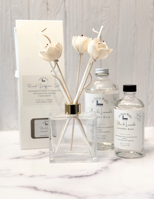 Lavender Rain Scented Reed Diffuser set and collection showcasing the optional floral reeds along with the included white reeds and additional for purchase bottles of 8oz or 16oz fragrance refills.  The whole scent collection is sitting on a white marble table with a white wood background.