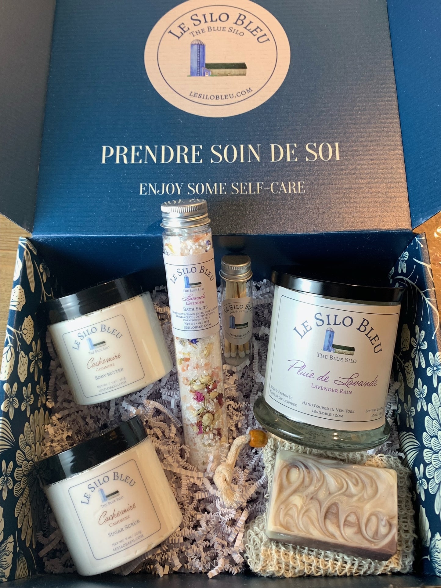 Le Silo Bleu branded premium gift box with soap, body butter, sugar scrub, a large 10oz candle, matches in a tube and a tube of bath salts all packaged in a premium branded box with blue and white flowers printed on it.