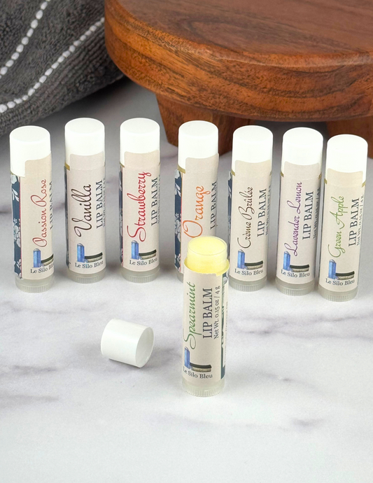 Lip Balm collection line up with one in front open all on a white marble table with a wood stand and grey towel in the background.