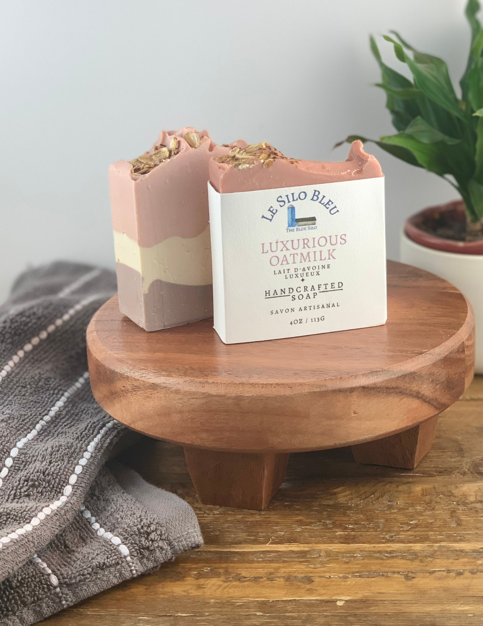 Two bars of Luxurious Oatmilk Soap sitting on a wooden platform with one soap wrapped in white labeling and one without. There is a green plant in a white pot in the background and a grey and white striped towel in the foreground. There is a wooden table and white background.
