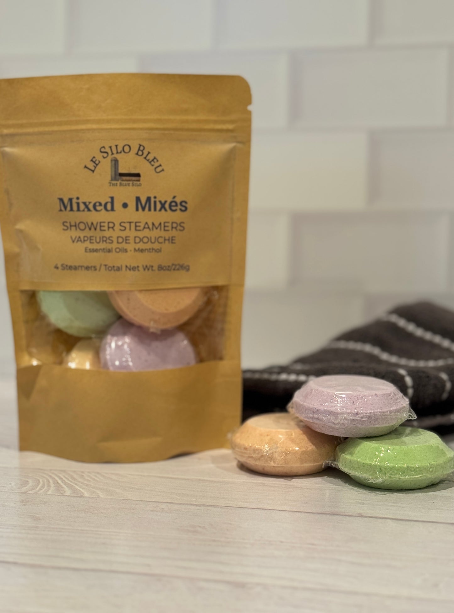 Mixed shower steamers in a brown craft bag sitting on a white wood table with a white tile background. There are three steamers and a grey towel with white stripes sitting on the table as well.