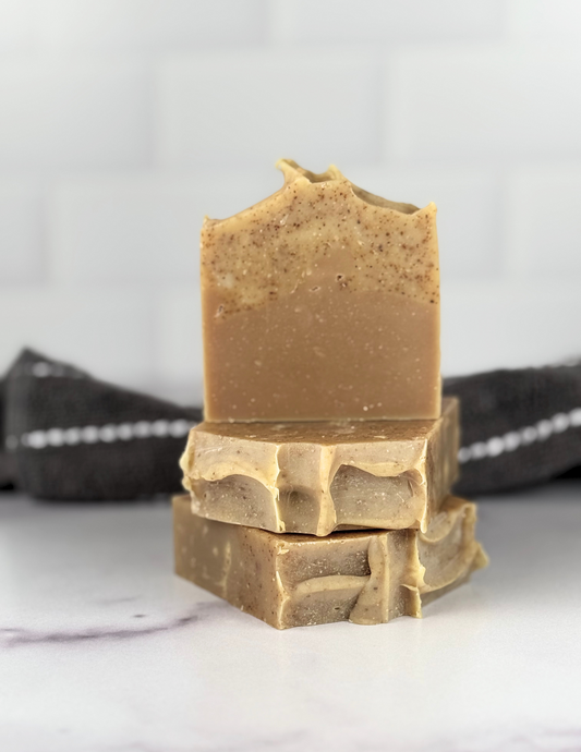 Stack of Mountain Man exfoliating soap on a white marble table with a grey striped towel in the back ground