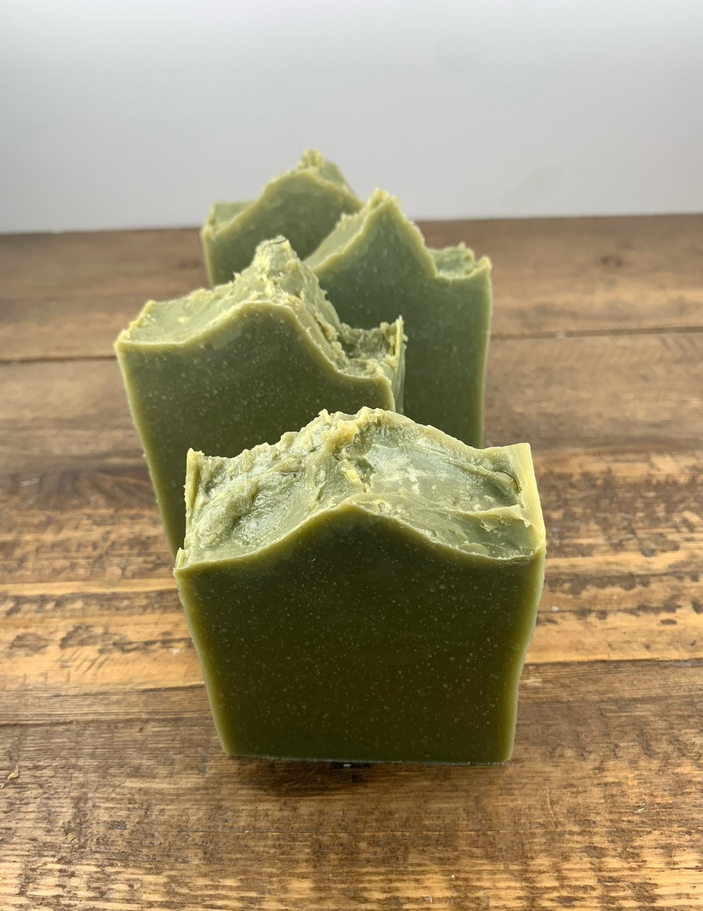 Four bars of this refreshing spearmint scented soap sit in a line atop a wooden table. This soap is a dark green from the spurlina powder and features essential oils.