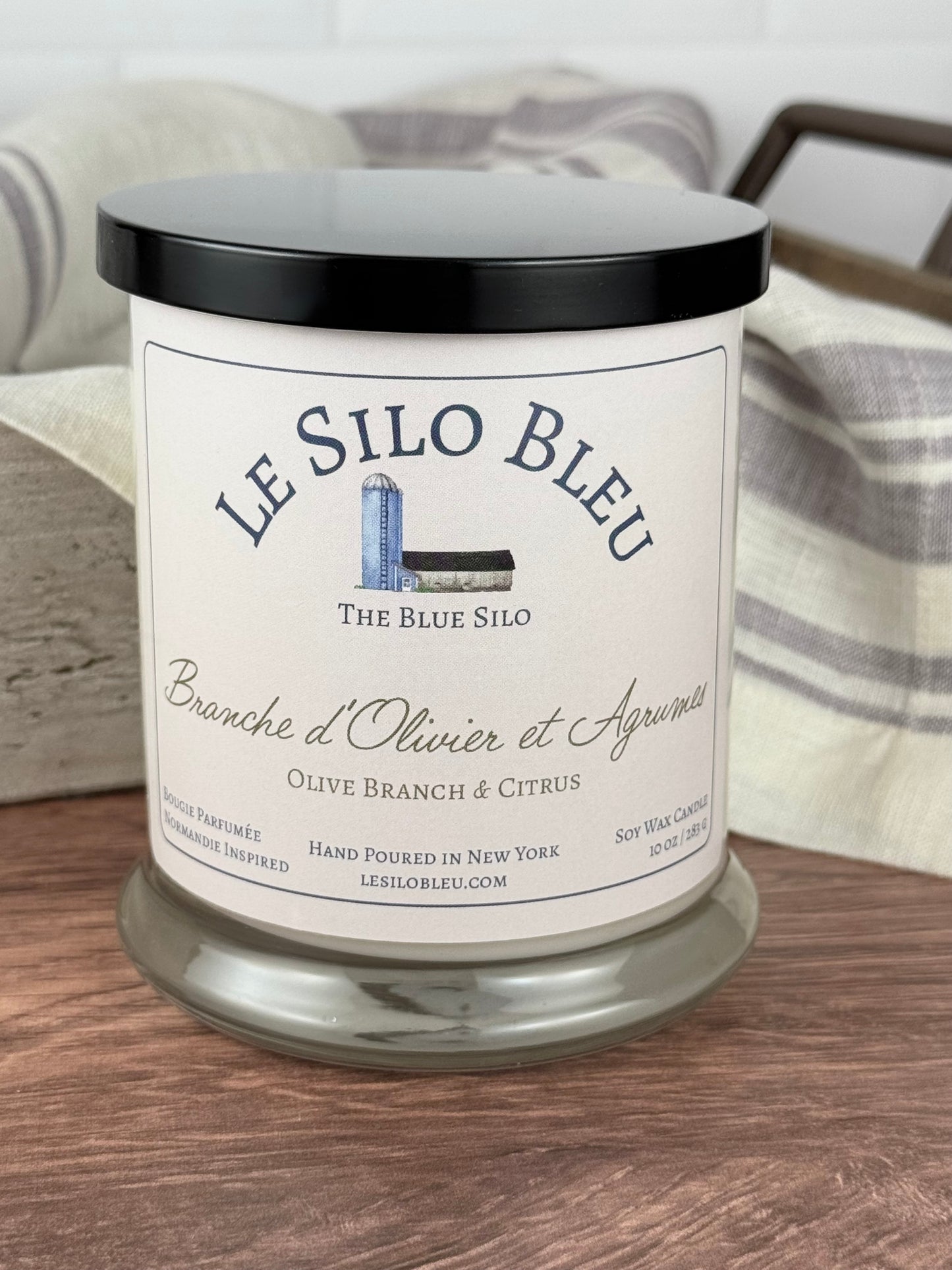 A large handpoured unlit soy wax candle in a glass jar labeled "Branche d'Olivier et Agrumes- Olive Branch and Citrus" sitting on a wooden table top with a black lid on the jar. In the background there is a white linen towel with lavender stripes in an antique box with a metal handle.