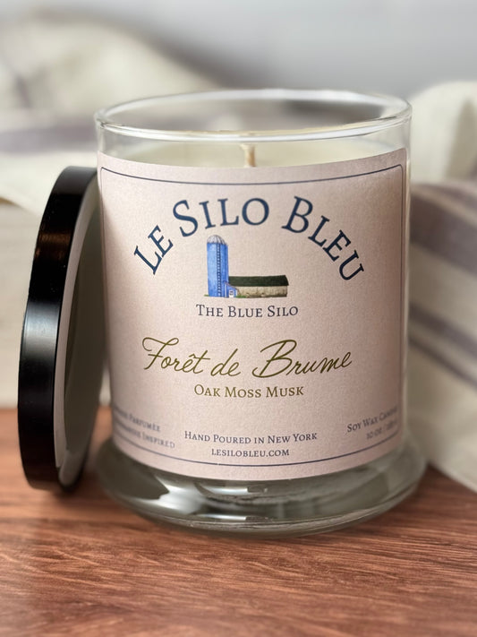 A large hand poured unlit soy wax candle in a glass jar labeled "Foret de Brume - Oak Moss Musk" sitting on a wooden tabletop with a black lid leaning on the jar. In the background there is a white linen towel with lavender stripes in an antique box.