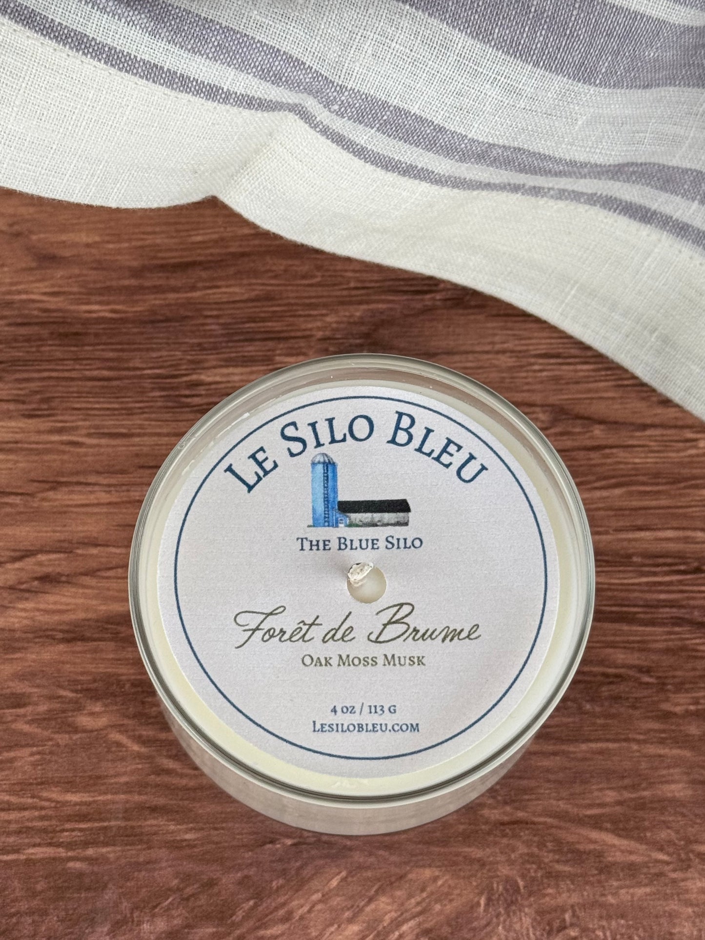 A small hand poured unlit soy wax candle in a glass jar labeled "Foret de Brume - Oak Moss Musk" sitting on a wooden tabletop In the background there is a white linen towel with lavender stripes in an antique box.