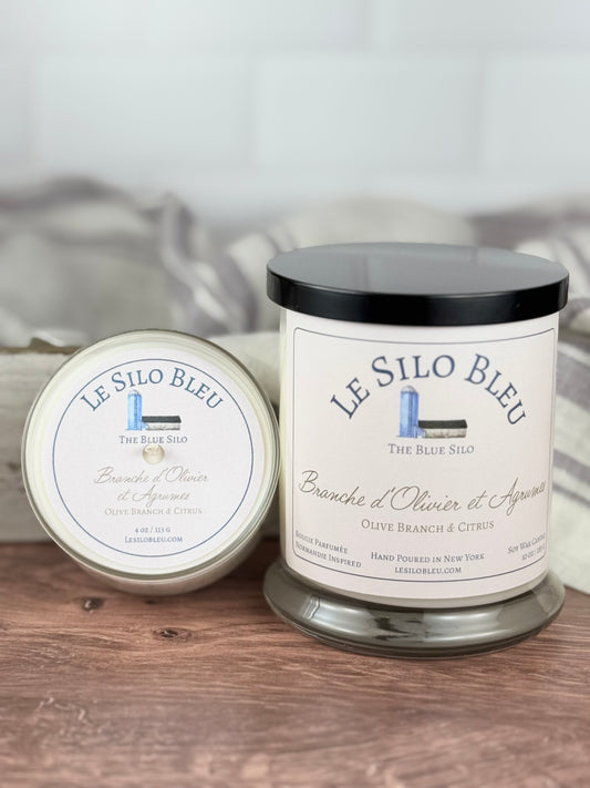 A large and small handpoured unlit soy wax candle in a glass jar labeled "Branche d'Olivier et Agrumes- Olive Branch and Citrus" sitting on a wooden table top with a black lid on the large jar. In the background there is a white linen towel with lavender stripes in an antique box with a metal handle.