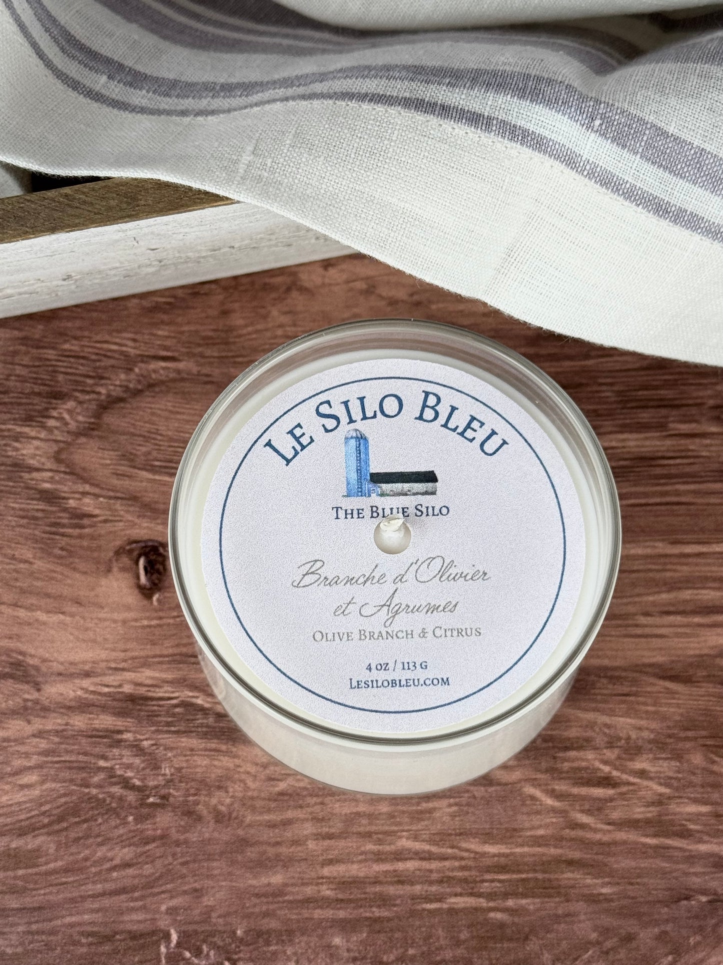 A small handpoured unlit soy wax candle in a glass jar labeled "Branche d'Olivier et Agrumes- Olive Branch and Citrus" sitting on a wooden table top. In the background there is a white linen towel with lavender stripes in an antique box with a metal handle.