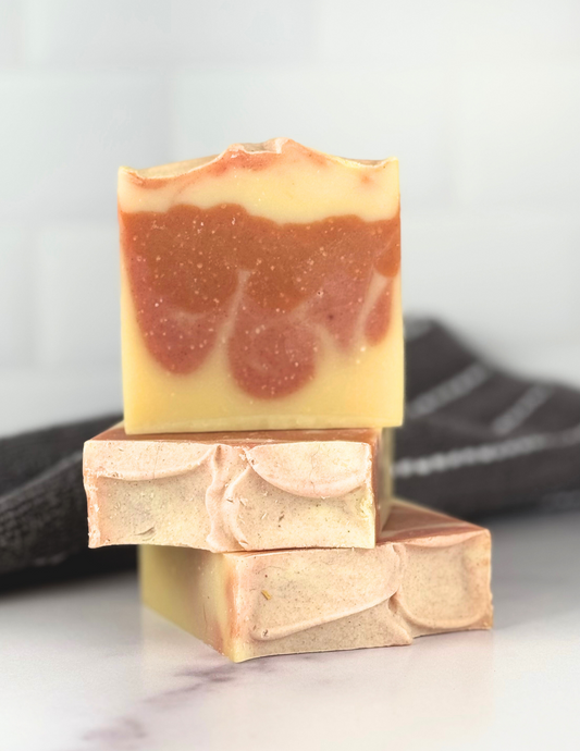 Three bars of Orange Blossom Soap sitting on a marble tabletop with a grey and white stripped towel in the background. The soap is two shades of orange and yellow swirled together.