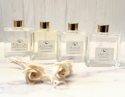 Scented bottles of diffuser fragrance collection and floral reeds along with the included white reeds is sitting on a white marble table with a white wood background.