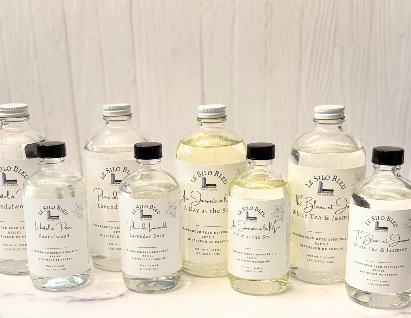 Reed Diffuser collection of scented refills that come in 8oz and 16oz bottles. They are all sitting on a white marble table with a white wood background.