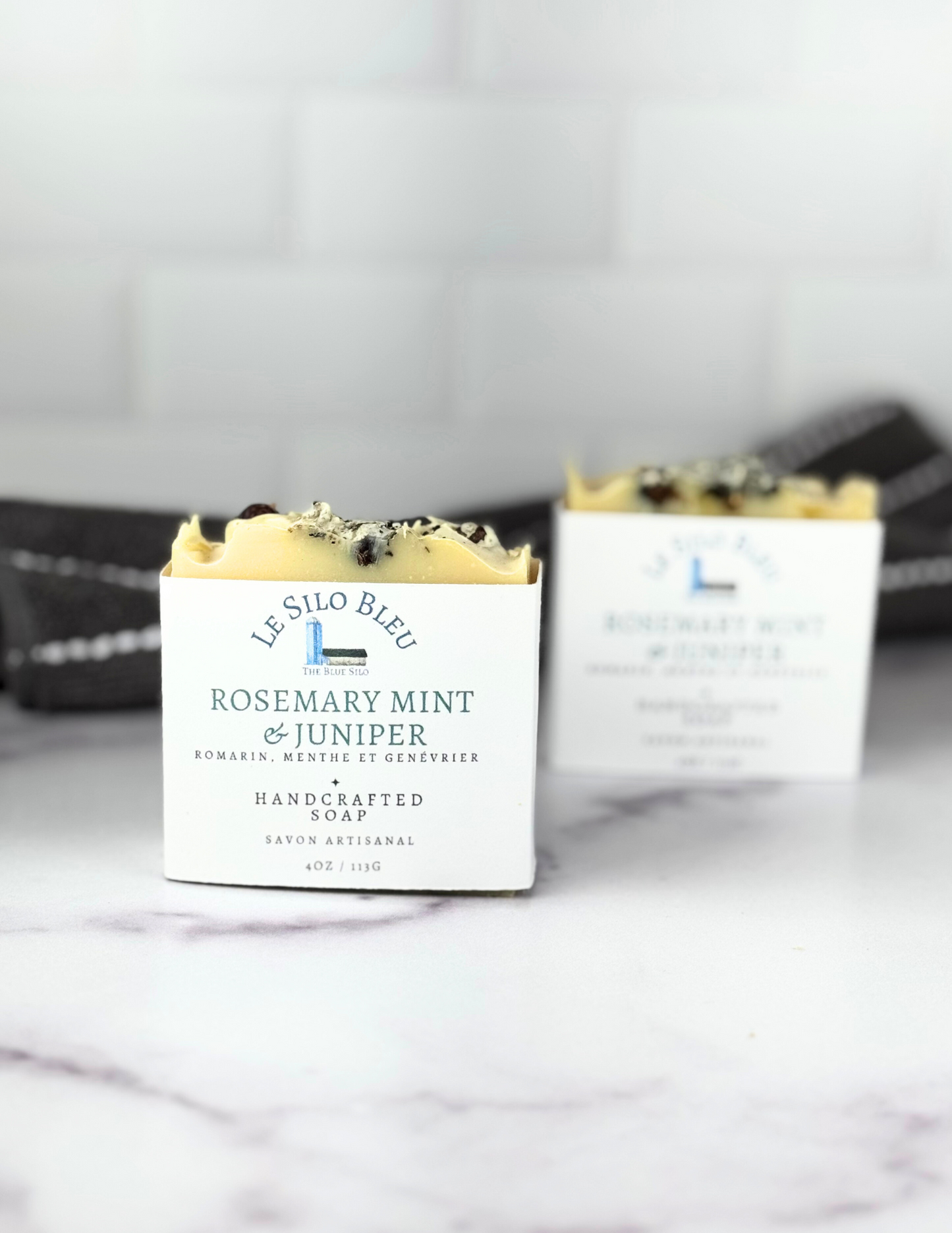 Two bars of labeled soap sit on a white marble tabletop with a grey and white stripped towel in the background.
