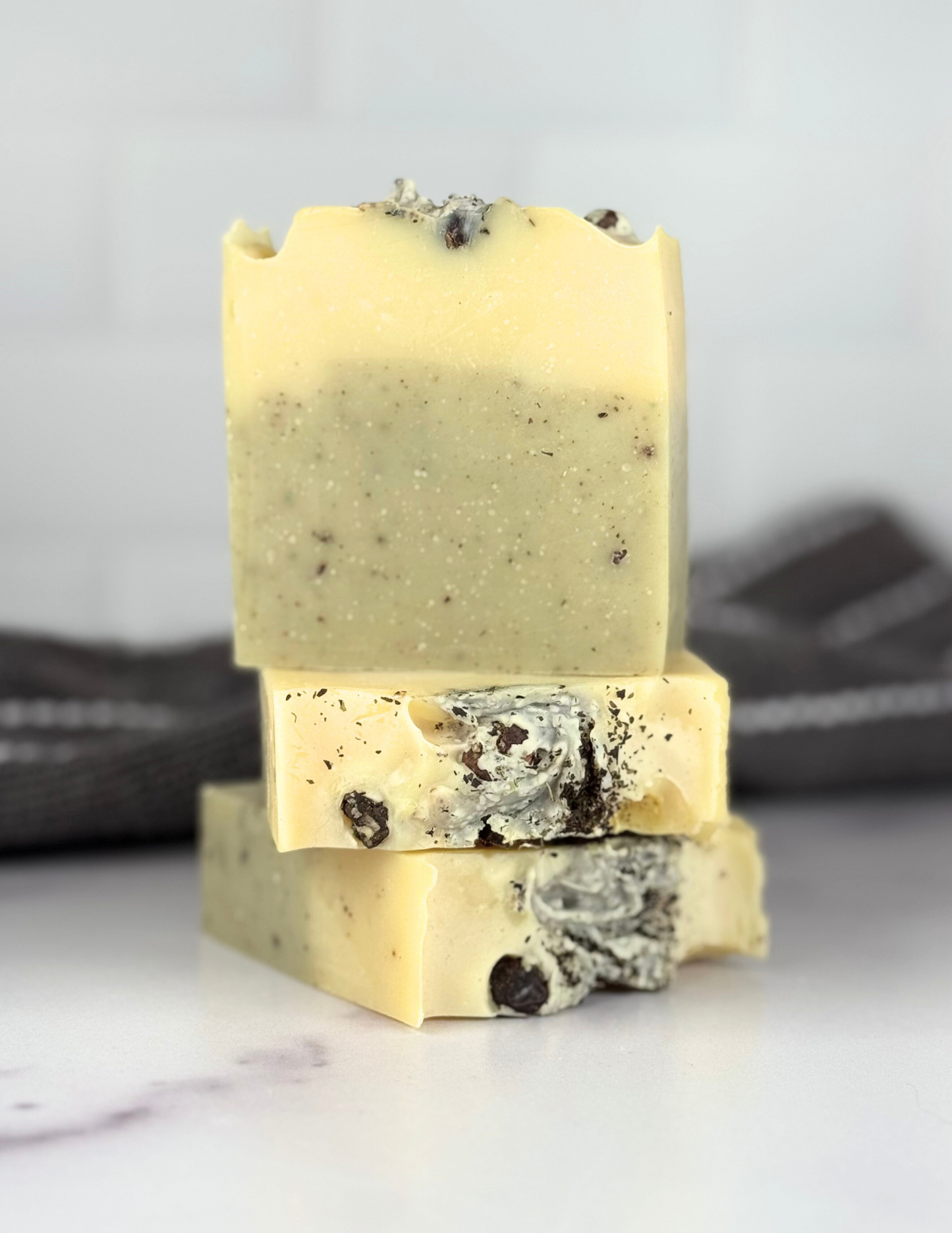 Three Bars of Rosemary Mint and Juniper Berry Soap sitting on a white marble tabletop with a grey and white stripped towel in the background. The soap is green and yellow and speckled with black and brown herbaceous pieces. The top has juniper berries visible.