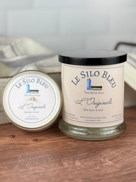 Twp sizes of hand poured unlit soy wax candles in a glass jars labeled " L'Originale-Sea Salt & Lily " sitting on a wooden tabletop with a black lid on the jar. In the background there is a white linen towel with lavender stripes in an antique box with a metal handle.