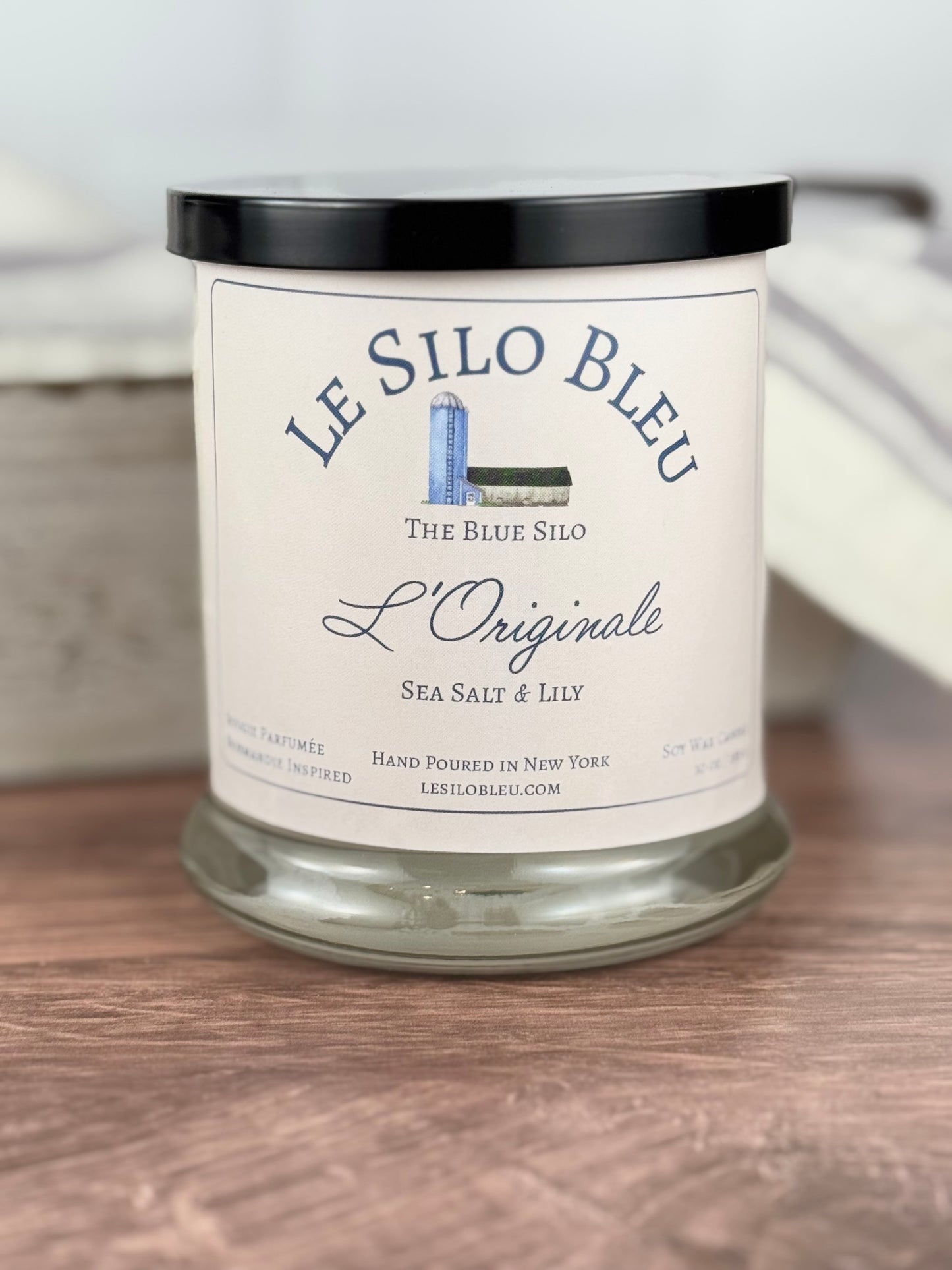 A handpoured unlit soy wax candle in a glass jar labeled " L'Originale Sea Salt Lily" sitting on a wooden table top with a black lid on the jar. In the background there is a white linen towel with lavender stripes in an antique box with a metal handle.
