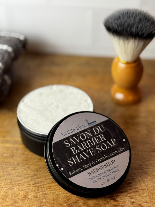 An open round tin of Le Silo Bleu Savon du Barbier Shave Soap sits on a wooden surface, with the lid placed in front. A shaving brush with dark bristles and a light wood handle stands blurred in the background, along with a folded towel.