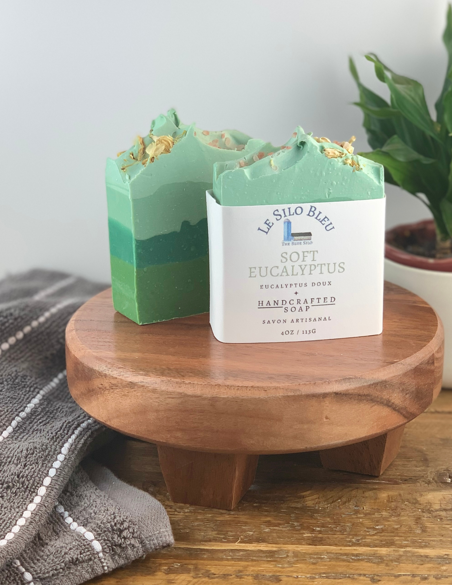 Two bars of Soft Eucalyptus Soap sitting on a wooden platform with one soap wrapped in white labeling and one without. There is a green plant in a white pot in the background and a grey and white striped towel in the foreground. There is a wooden table and white background.