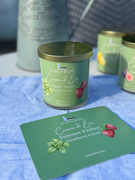 Summer Kitchen Strawberry & Basil Candle with a green label on a blue surface, surrounded by similar candles and a pitcher of flowers in the background.