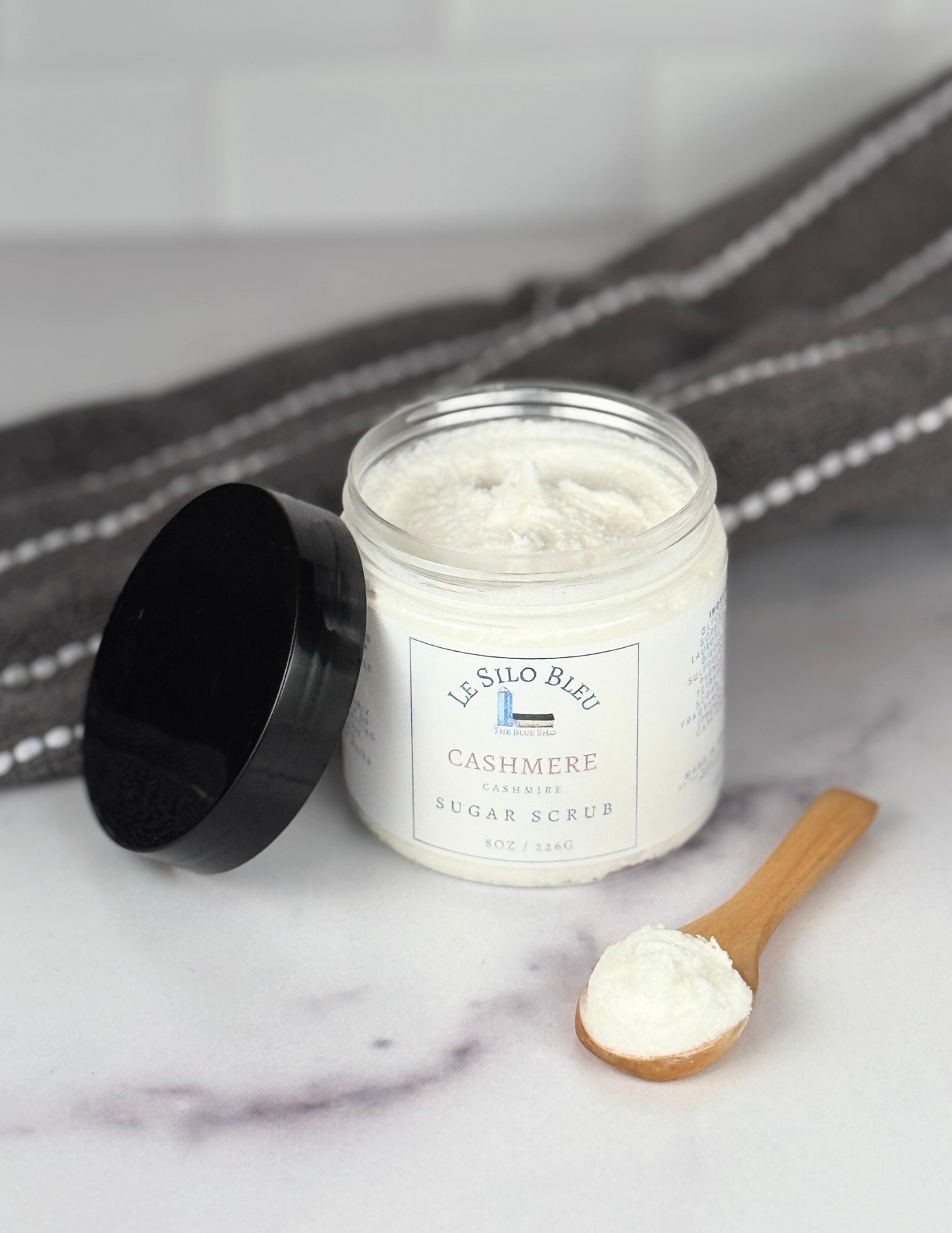 Jar of sugar scrub jar with black lid leaning against the jar. Both are sitting on a white marble table with a grey and white stripped towel in the background. The white labels for these show the logos and names of the scent- Cashmere. There is also a small wooden spoon with a scoop of sugar scrub on it in the foreground.