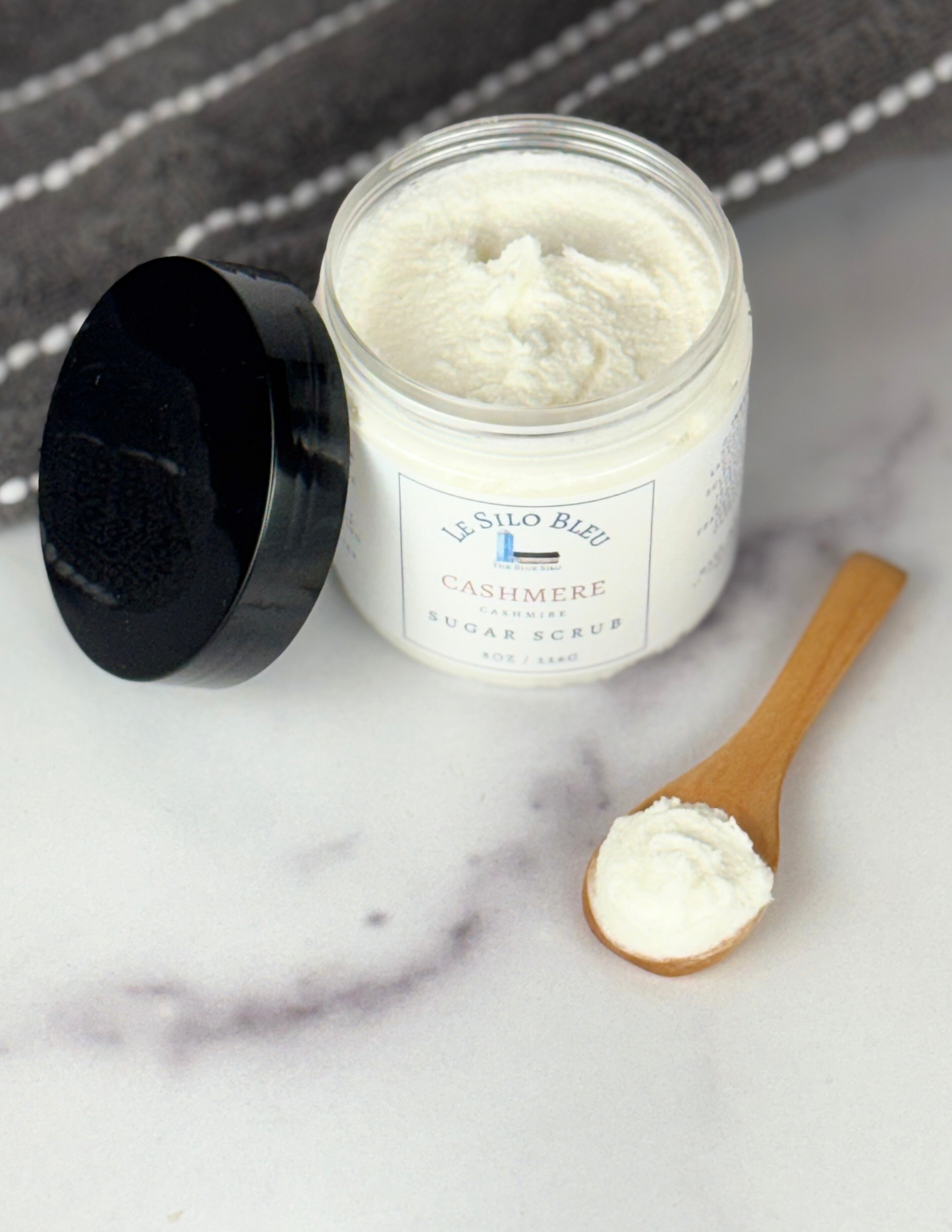 An open jar of Cashmere scented Sugar Scrub with a black lid leaning against the jar sitting on top of a white marble table with a grey and white stripped towel in the background. The label shows the logo and the name of the scent. There is a small wooden scoop spoon with sugar scrub in it on the table as well.