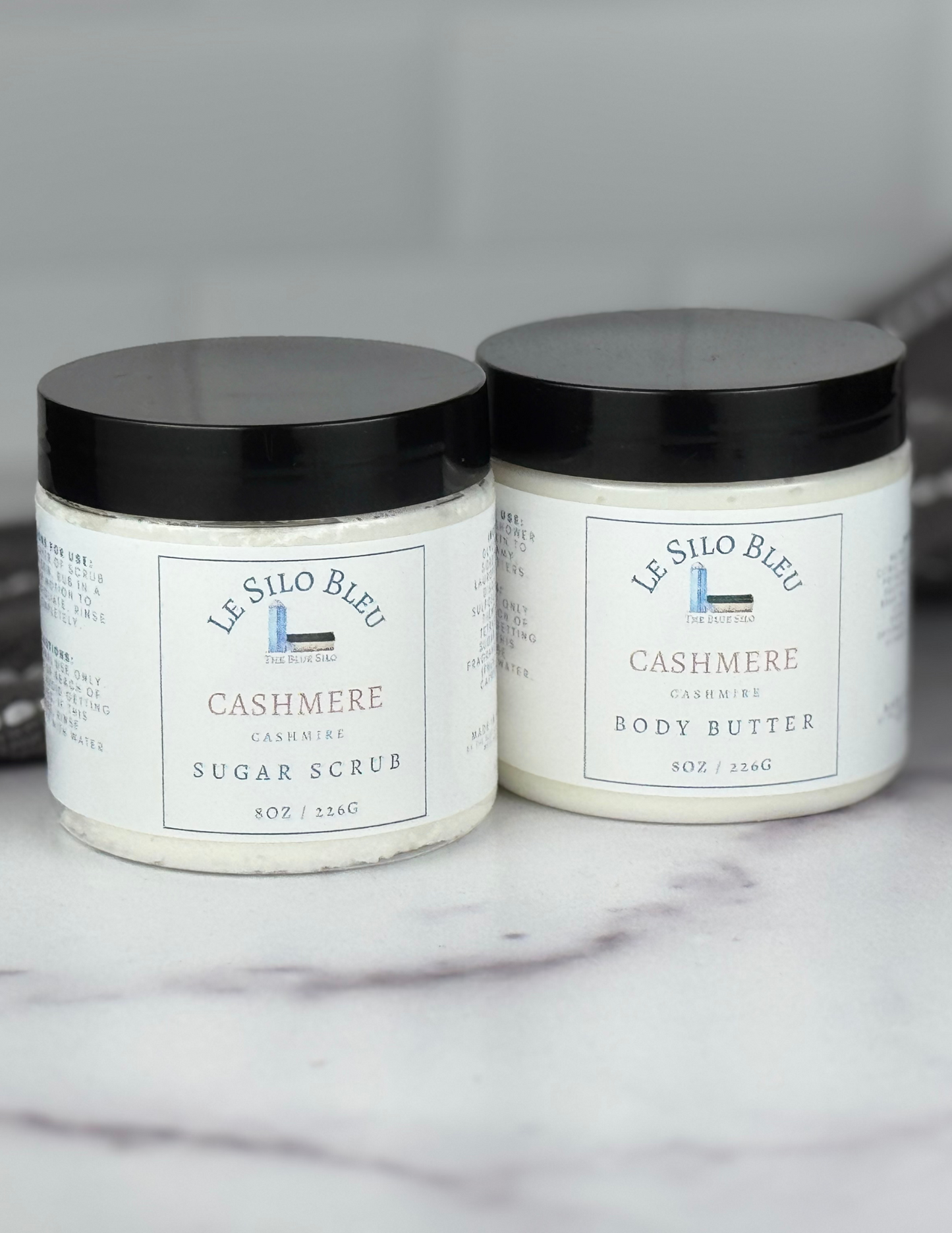 Set of body butter and sugar scrub jars with black lids sitting on a white marble table with a grey and white stripped towel in the background. The white labels for these show the logos and names of the scent- Cashmere.