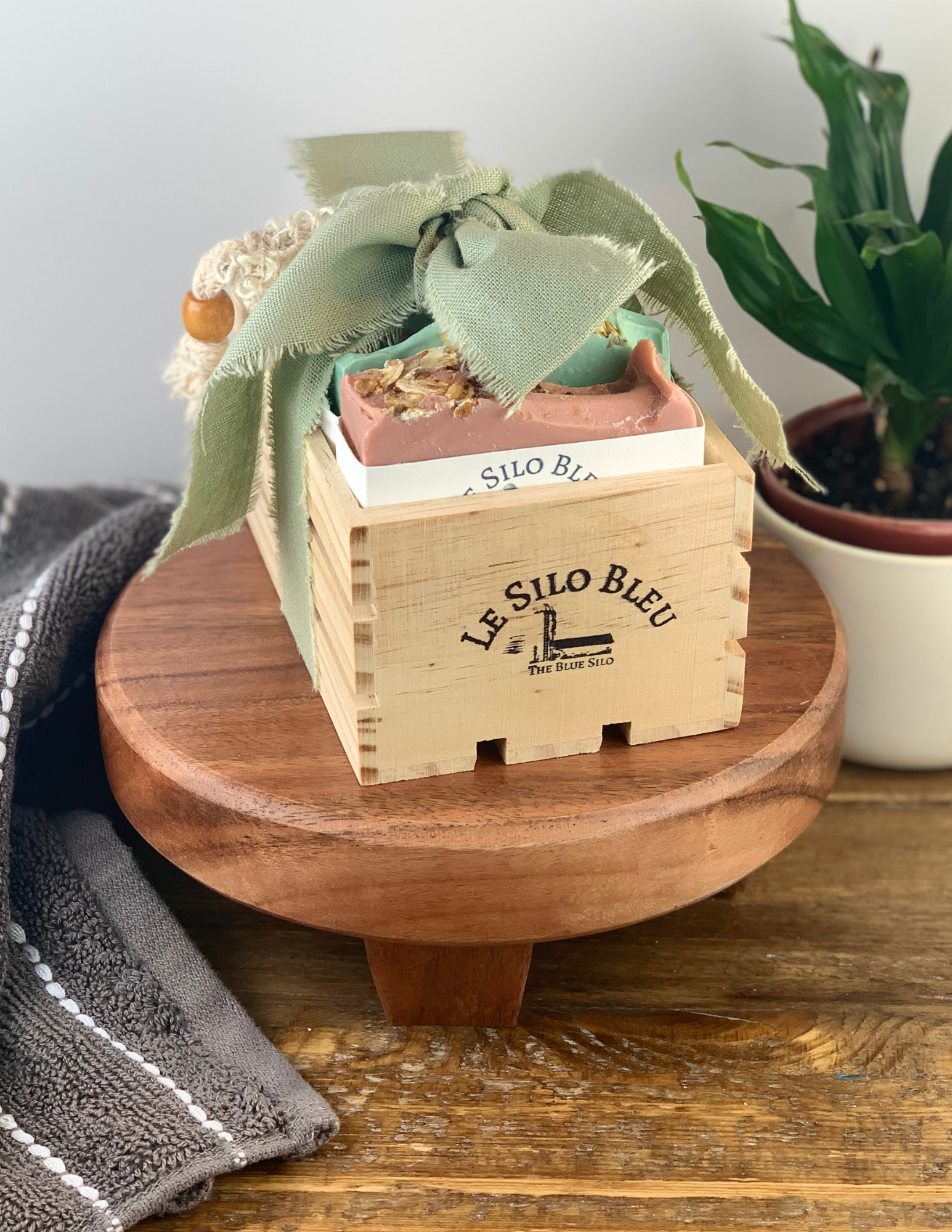 On a brown woodtopped table sits a wood stand with a small natural wood crate filled with three bars of our best selling soaps and an exfoliating scrub soap bag. This bundle is tied with a bow that is green and has the wooden box stamped with our logo. There is a green plant in a white pot in the background and a grey and white stipped hand towel on the table in the foreground. There is a white background and a wooden table top.