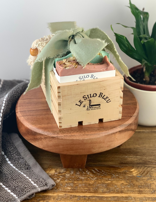 On a brown woodtopped table sits a wood stand with a small natural wood crate filled with three bars of our best selling soaps and an exfoliating scrub soap bag. This bundle is tied with a bow that is green and has the wooden box stamped with our logo. There is a green plant in a white pot in the background and a grey and white stipped hand towel on the table in the foreground. There is a white background and a wooden table top.